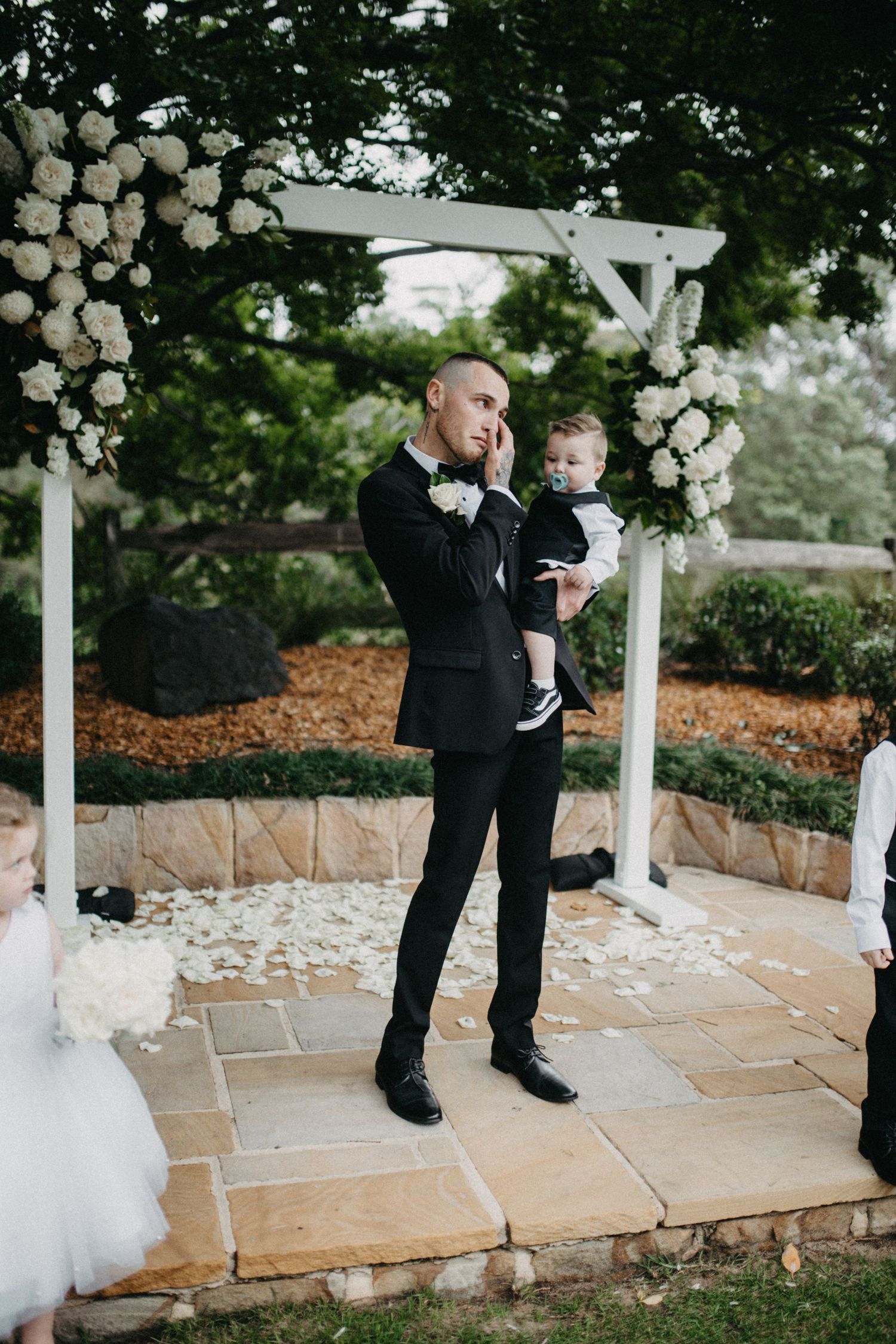Adorable wedding moment captured on stone patio with white floral arrangements and petals scattered around.