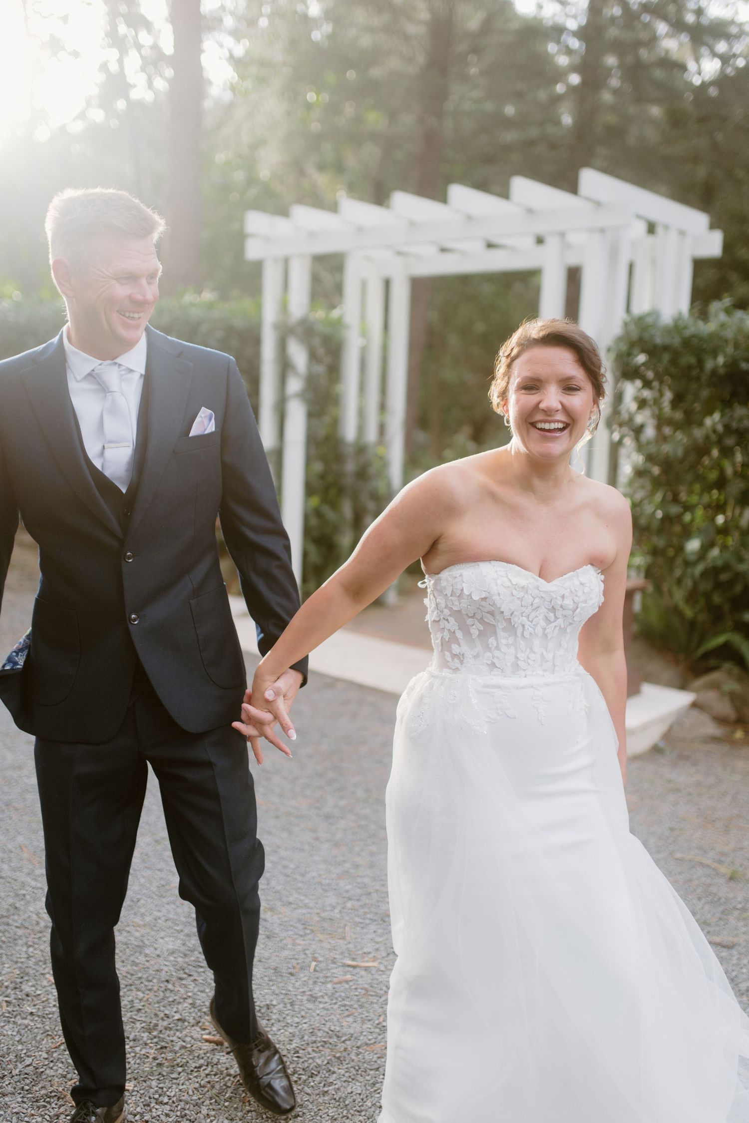 A bride in a strapless lace wedding dress walks joyfully with her groom on a gravel path.