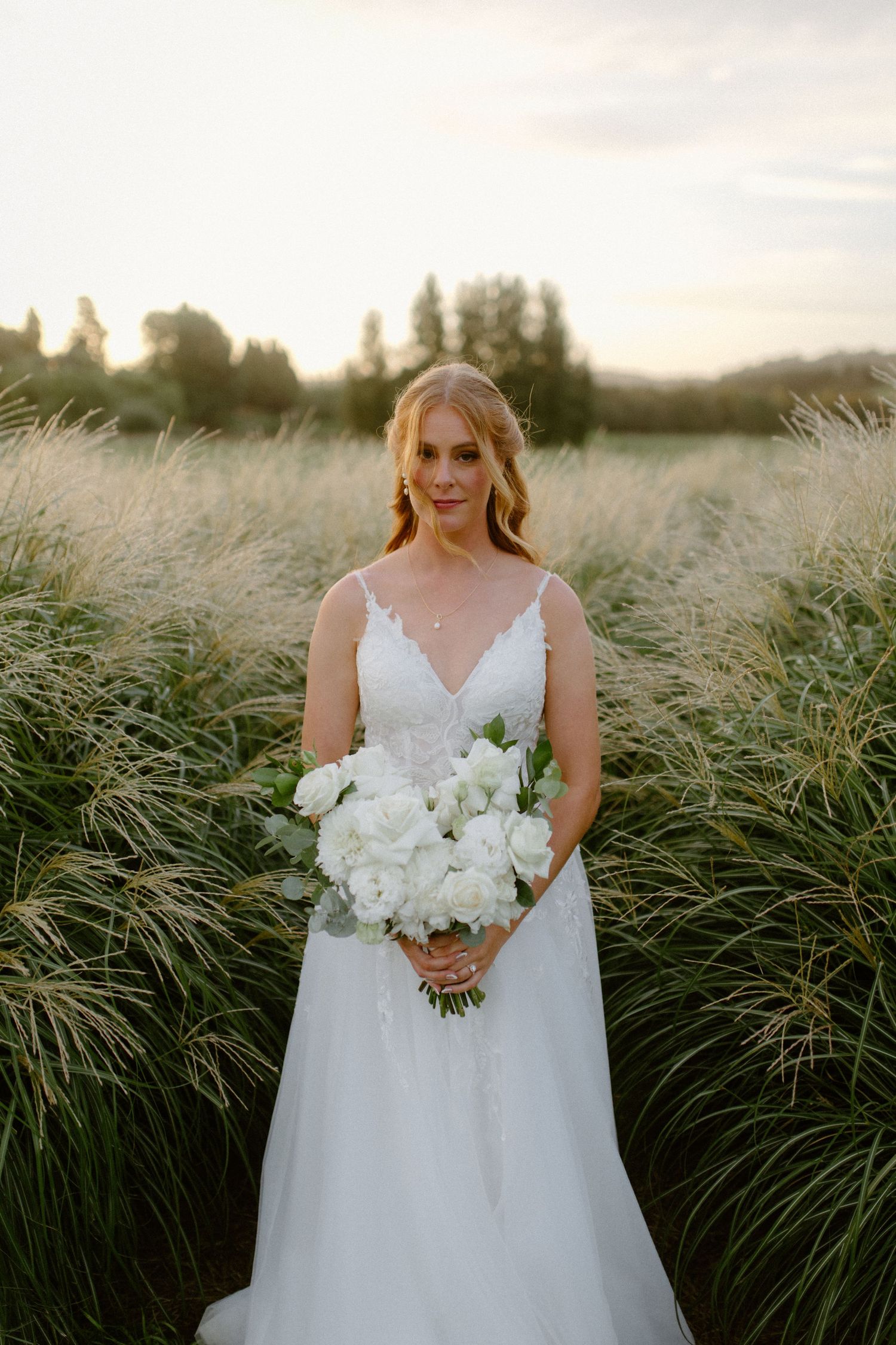 Bride in white gown holds elegant white bouquet while standing in tall grass at sunset.