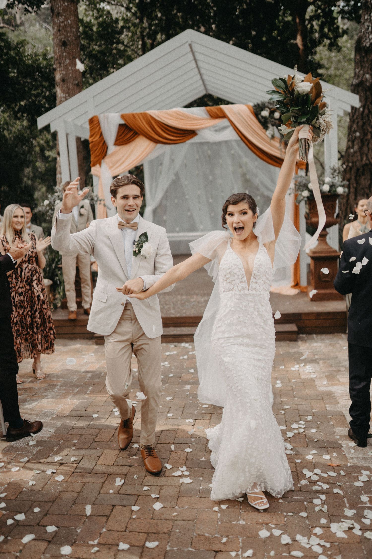 Lively wedding celebration with the couple dancing and twirling on a brick patio with flower petals scattered around.