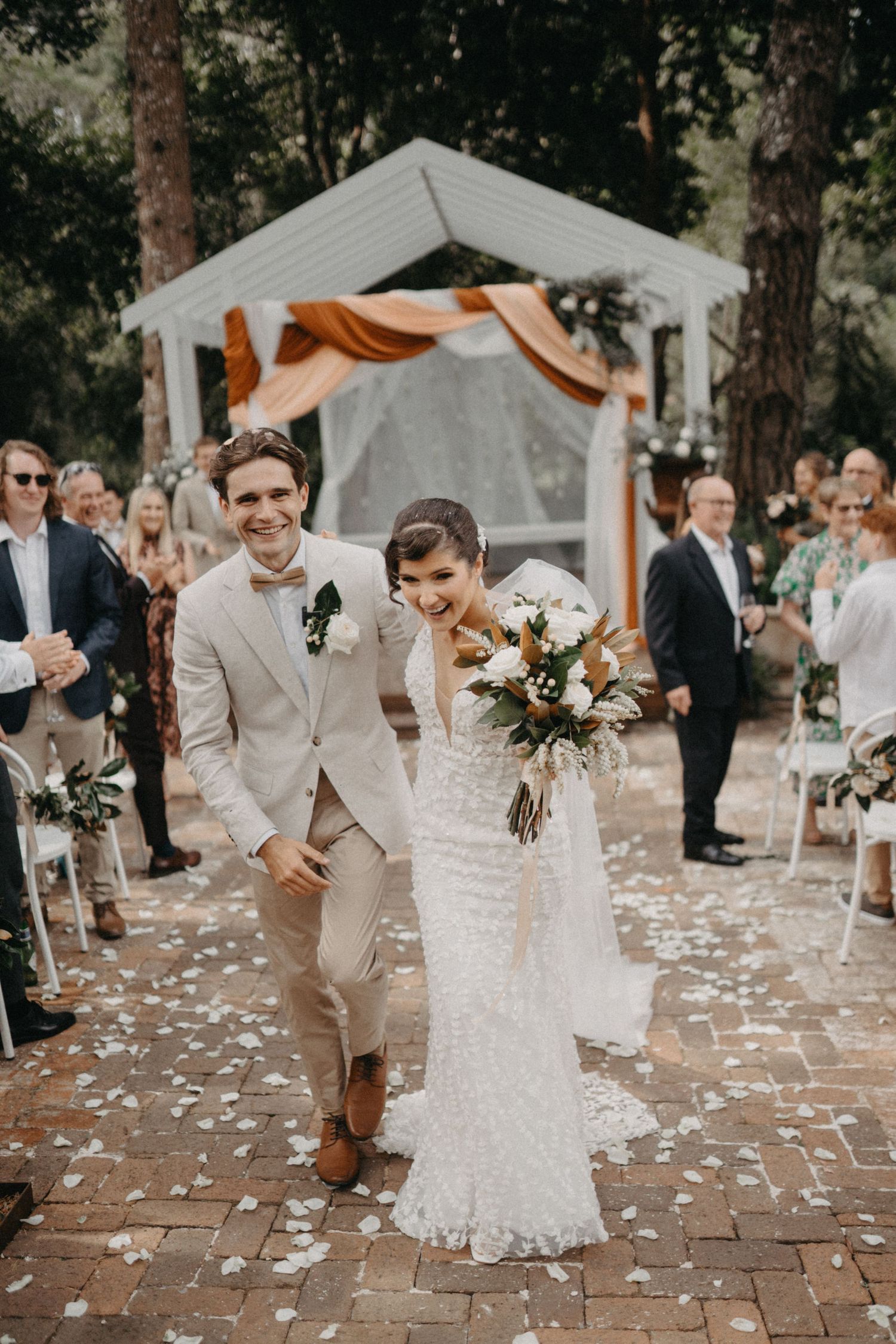 Wedding party dancing joyfully on a brick patio surrounded by trees with orange decorative fabric in the background.