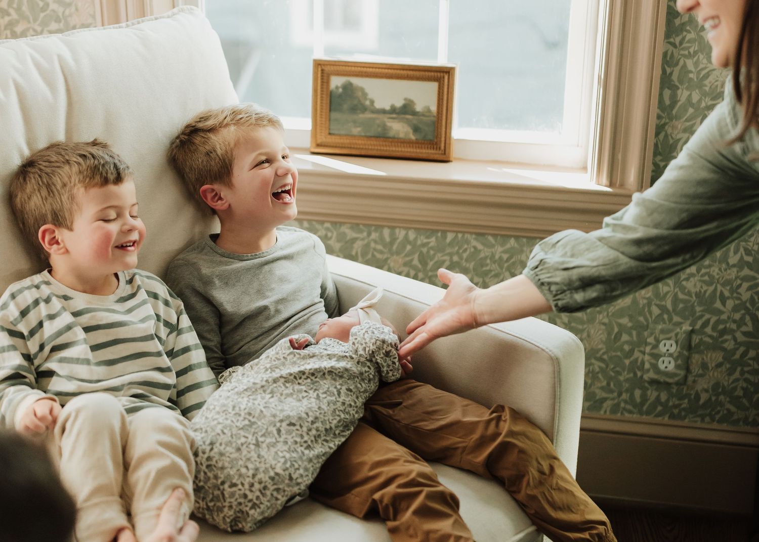 Two young children sit on a couch laughing together while someone reaches out to tickle them.