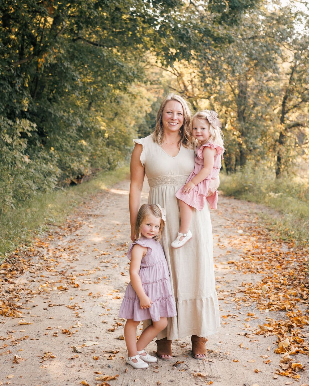 A family in neutral-toned dresses walks along a sunlit autumn path covered in fallen leaves.