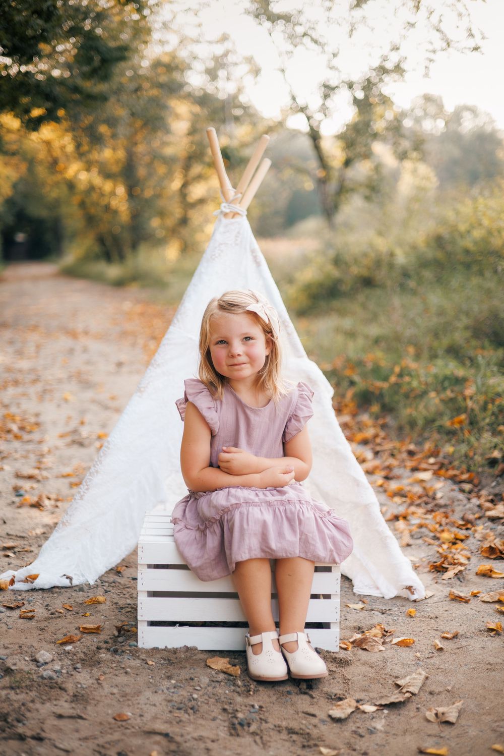 Young blonde girl in pink dress sitting on white crate before teepee tent in autumn forest setting.