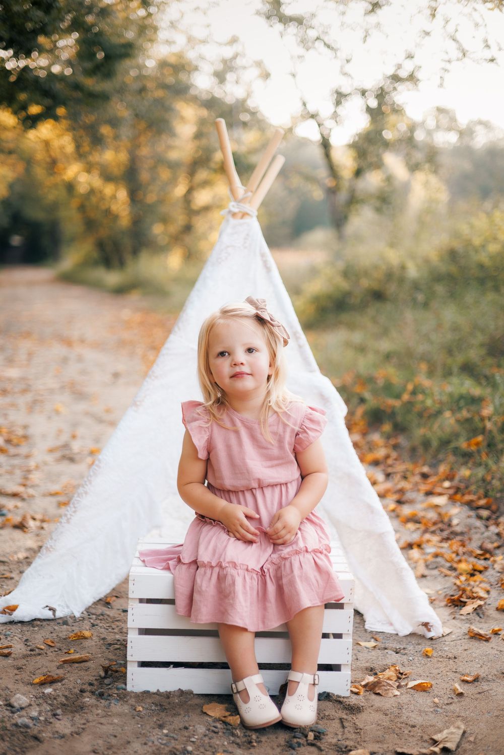 A young girl in a pink dress sits on a white wooden crate in front of a white teepee outdoors.