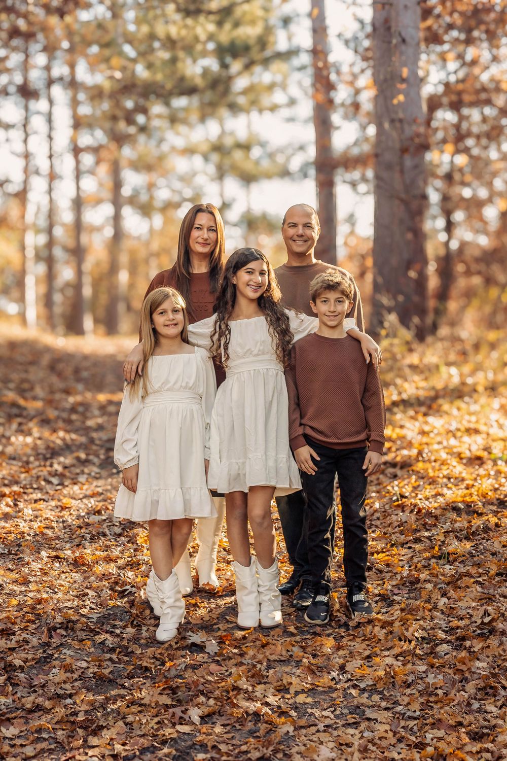Family of five posing in autumn forest with fallen leaves, two girls in white dresses and a boy with parents behind them.