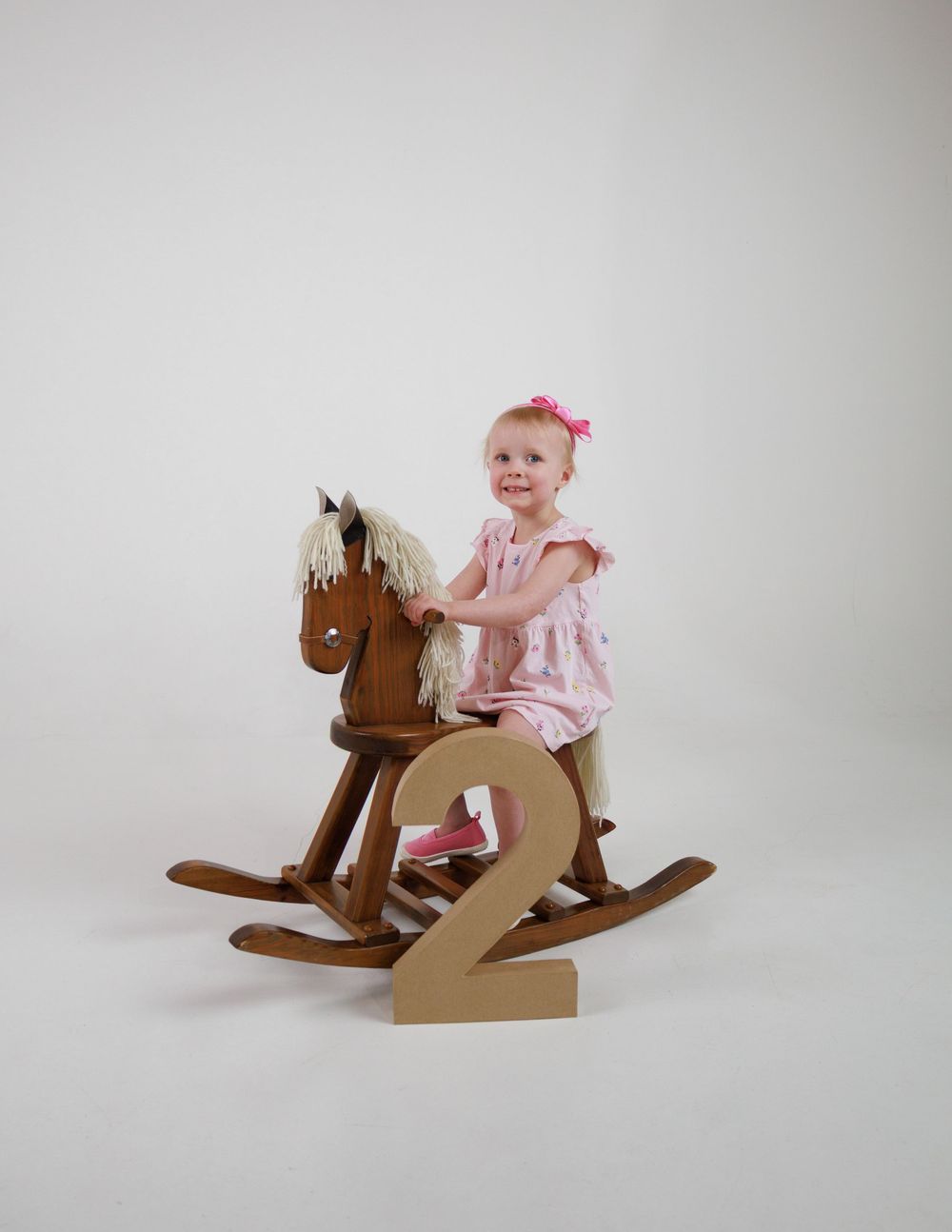 A smiling toddler girl in a pink dress sits on a wooden rocking horse next to a number 2, celebrating her 2nd birthday.