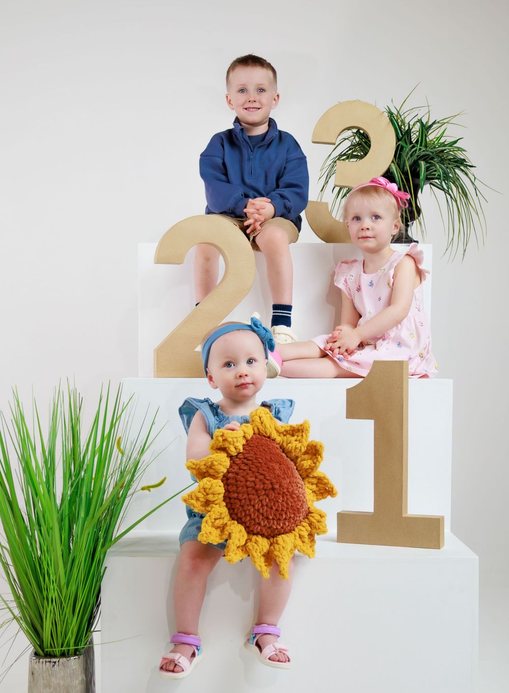 Three children pose on white podiums with gold numbers 2, 3, and 1, with the youngest wearing a crochet sunflower outfit.