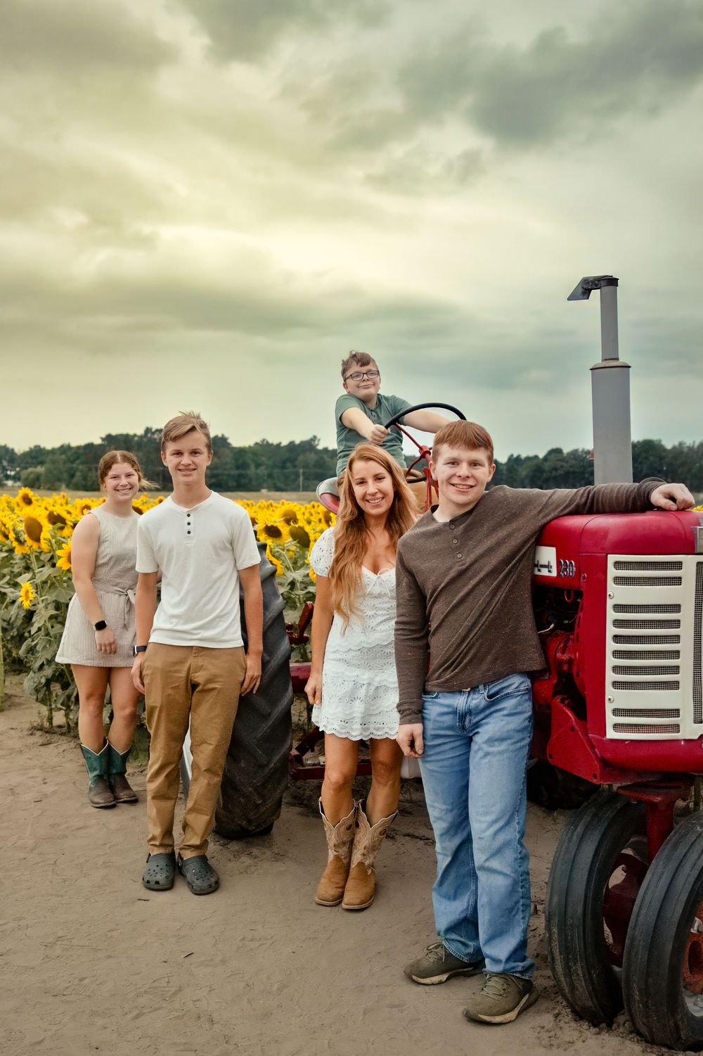 Family of five posing near a red tractor beside a sunflower field on a cloudy day.