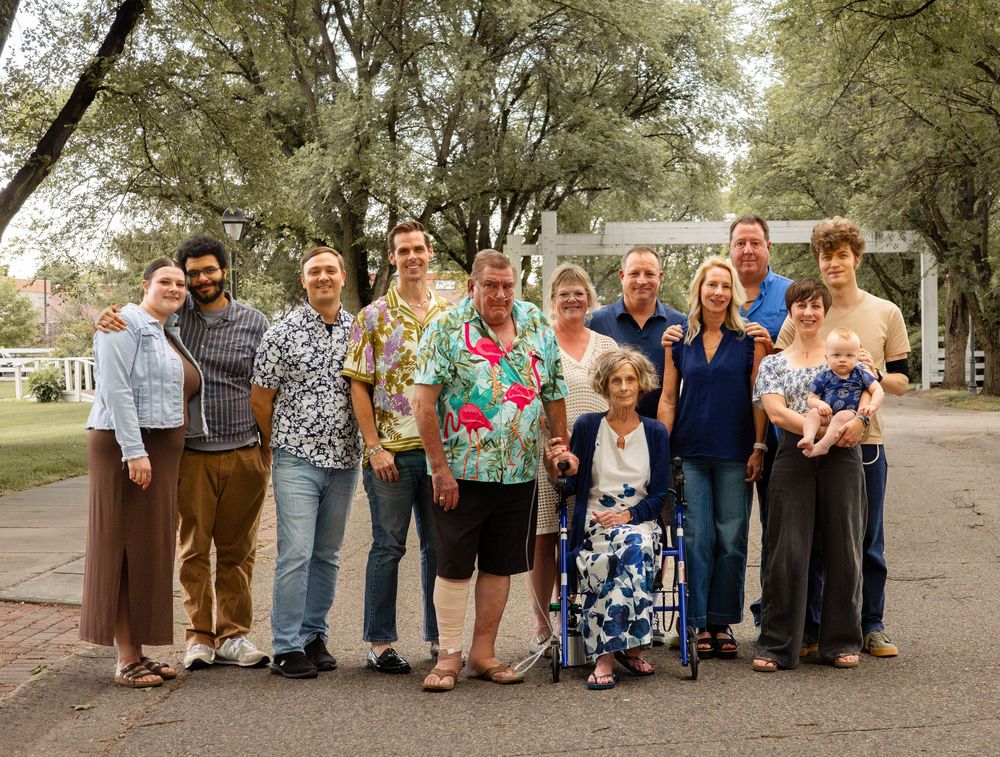 Large family group photo outdoors under shaded trees, smiling together on a sunny day.