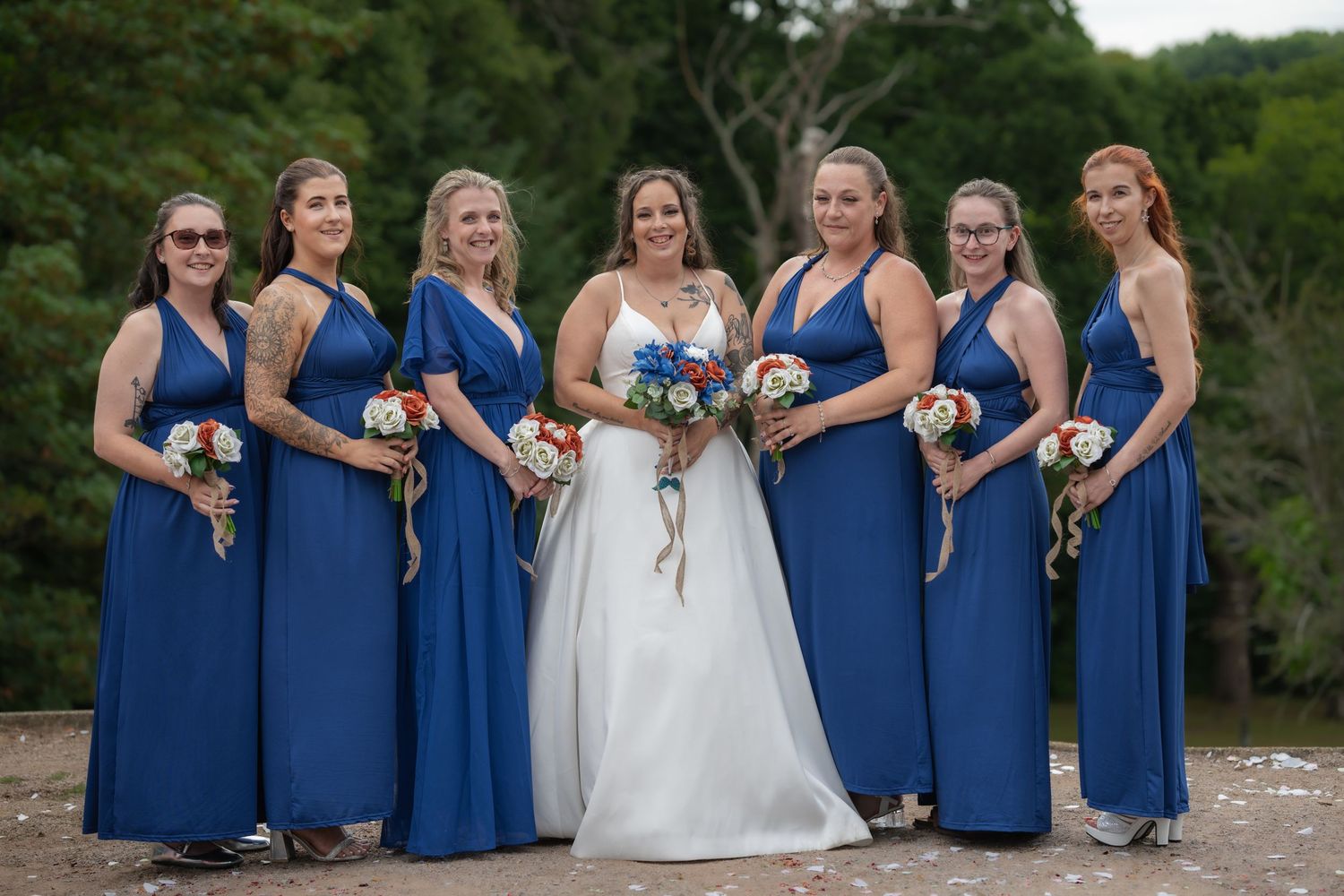Wedding party in matching royal blue floor-length dresses posing together outdoors in natural setting.