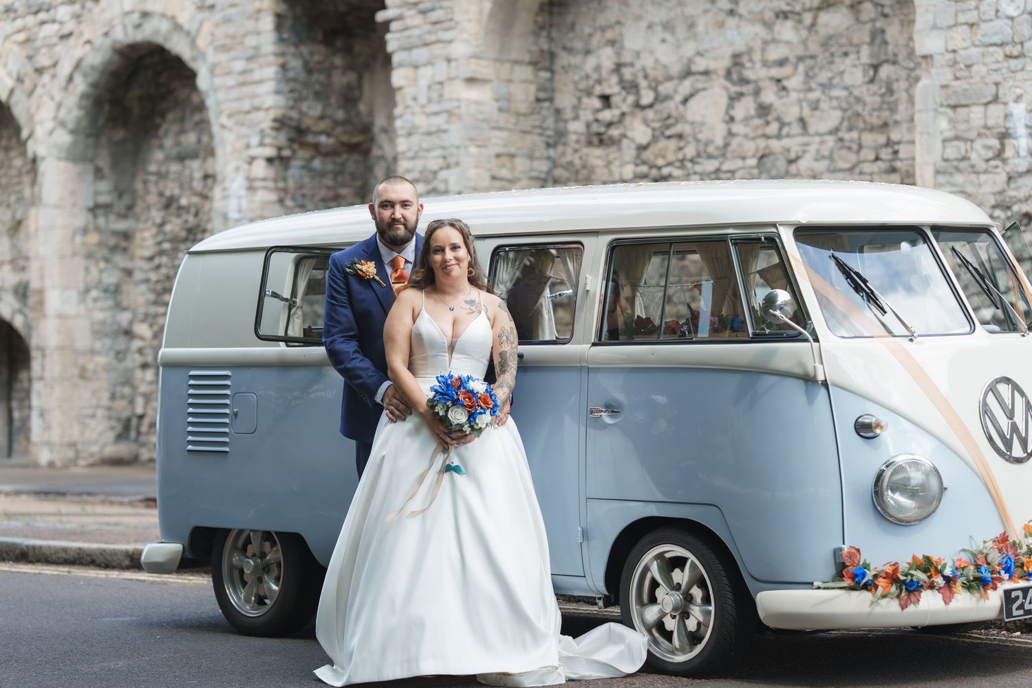 A light blue vintage Volkswagen van serves as a wedding vehicle parked in front of a stone castle wall.