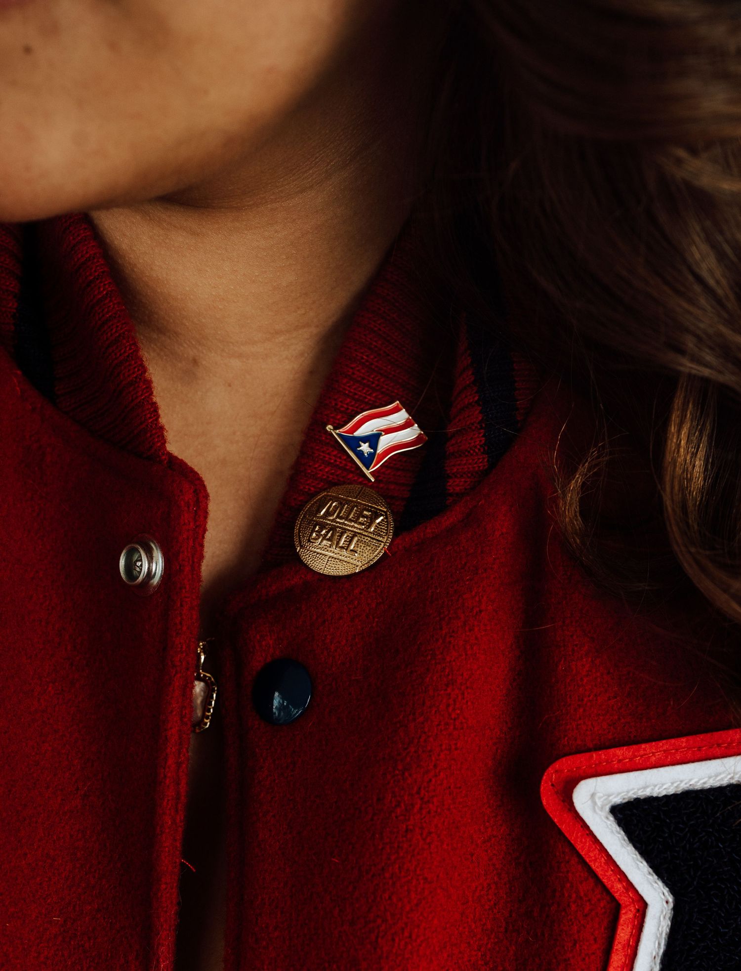 Close-up detail of a red volleyball varsity jacket with enamel pins including a Puerto Rican flag and other decorative buttons.