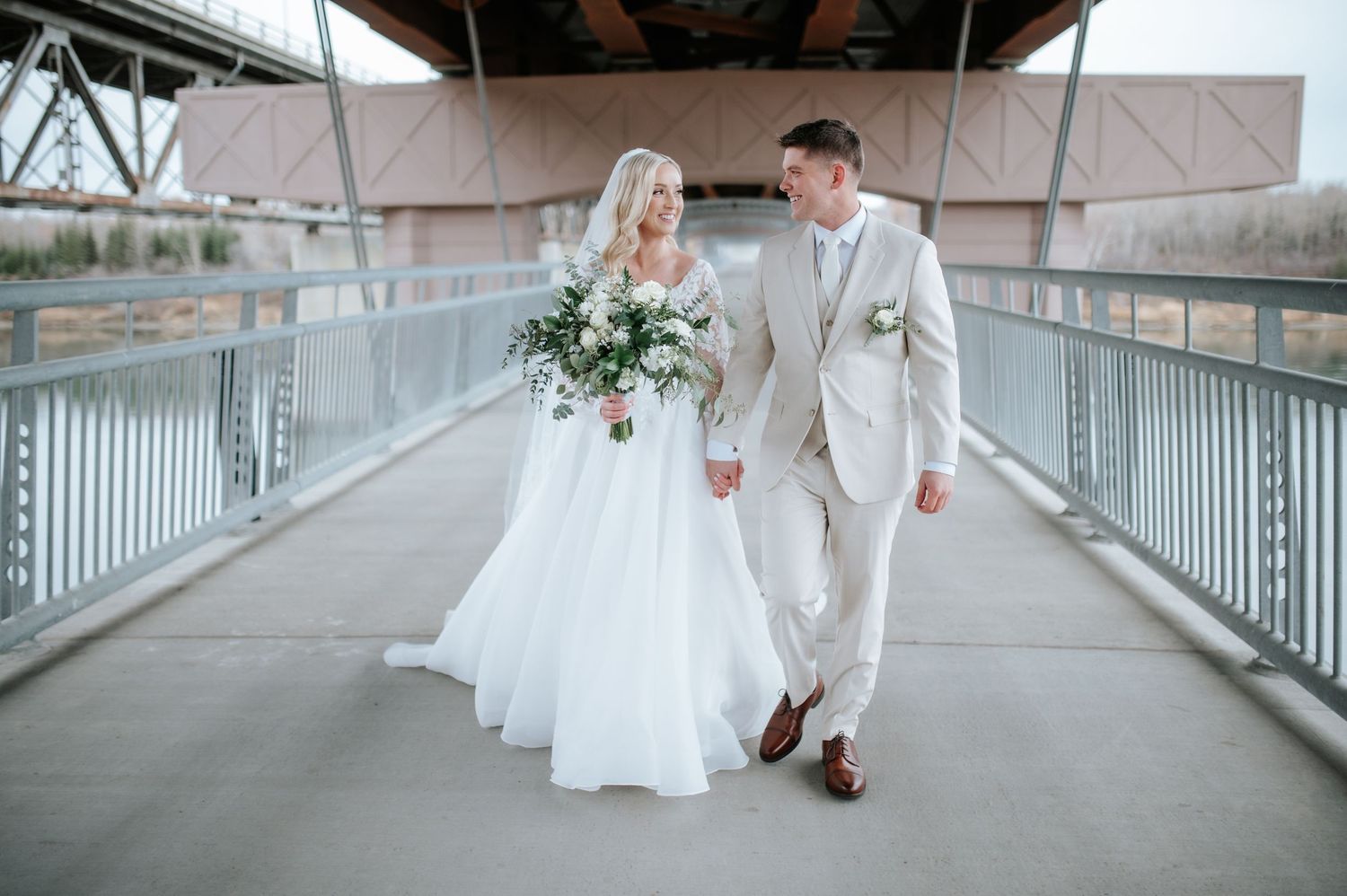 Wedding couple walking together on a modern bridge in light colored attire with a floral bouquet.