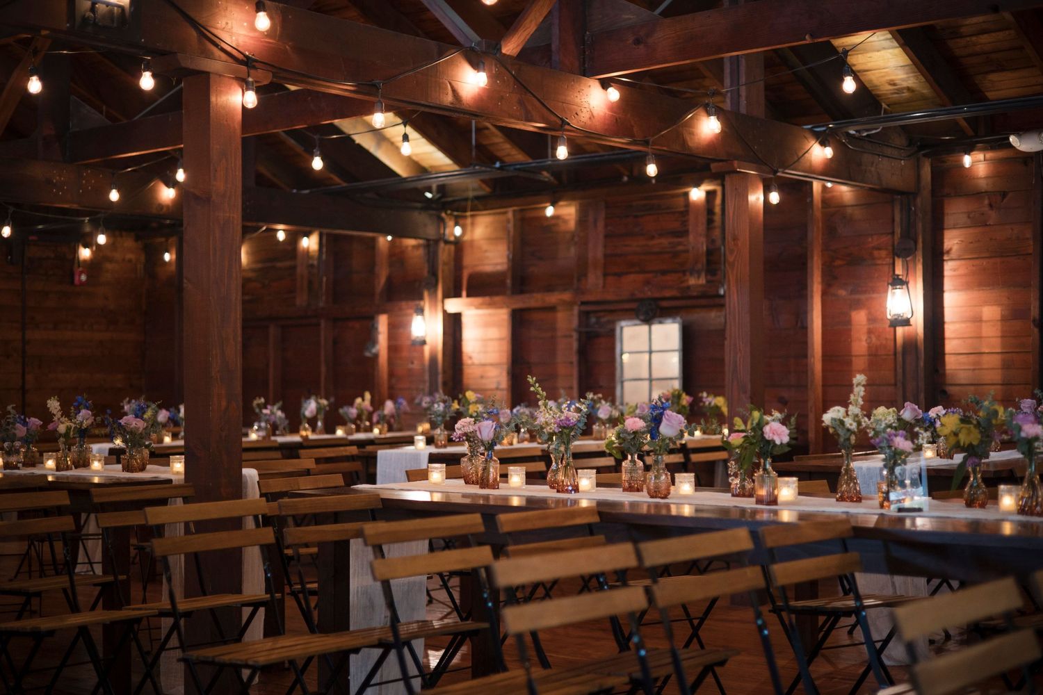 Interior of a rustic wooden event venue with warm lighting, long tables, and bench seating in banquet style setup.