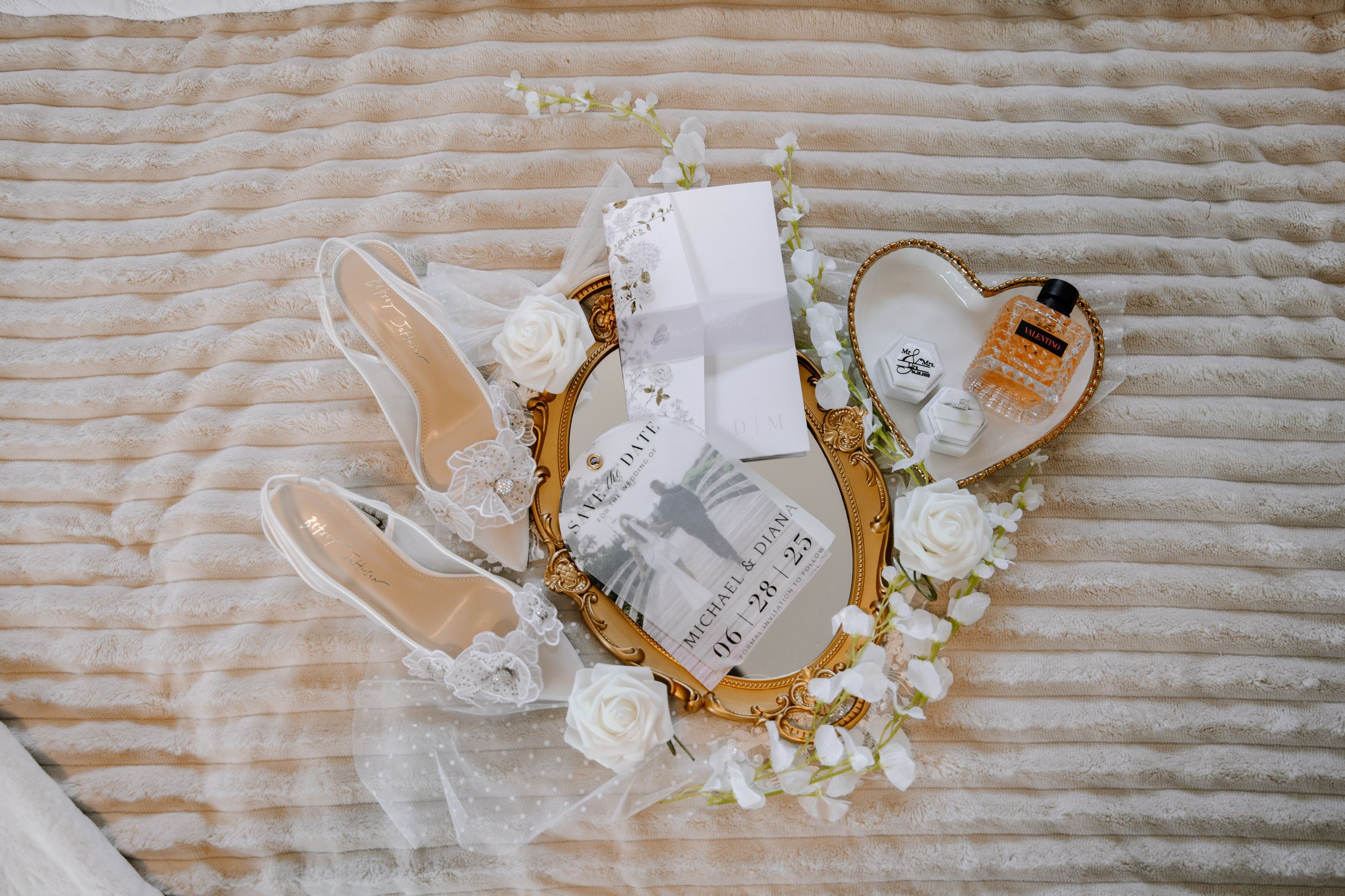 Clear and white wedding shoes with small floral details arranged on a beige quilted surface with scattered flower petals.