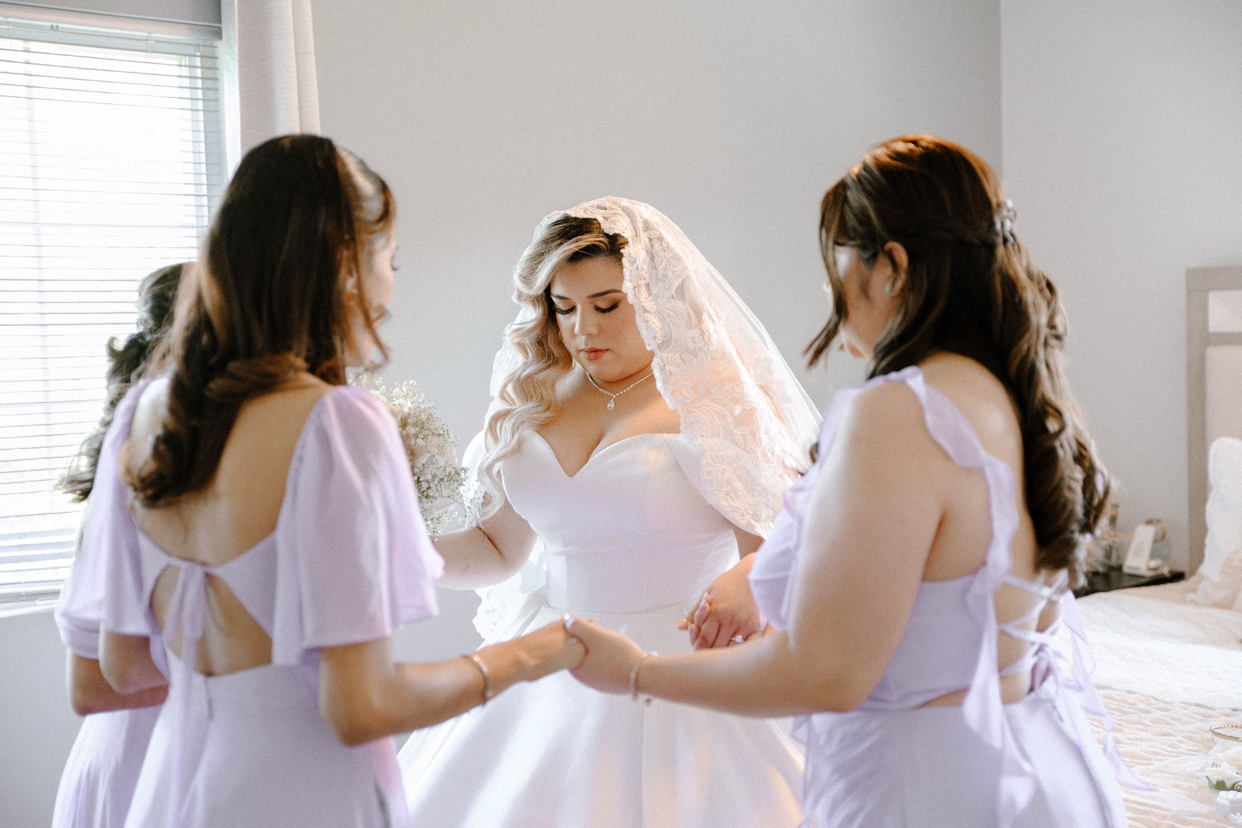 Bridesmaids in light colored dresses help the bride get ready in a bright sunlit room.
