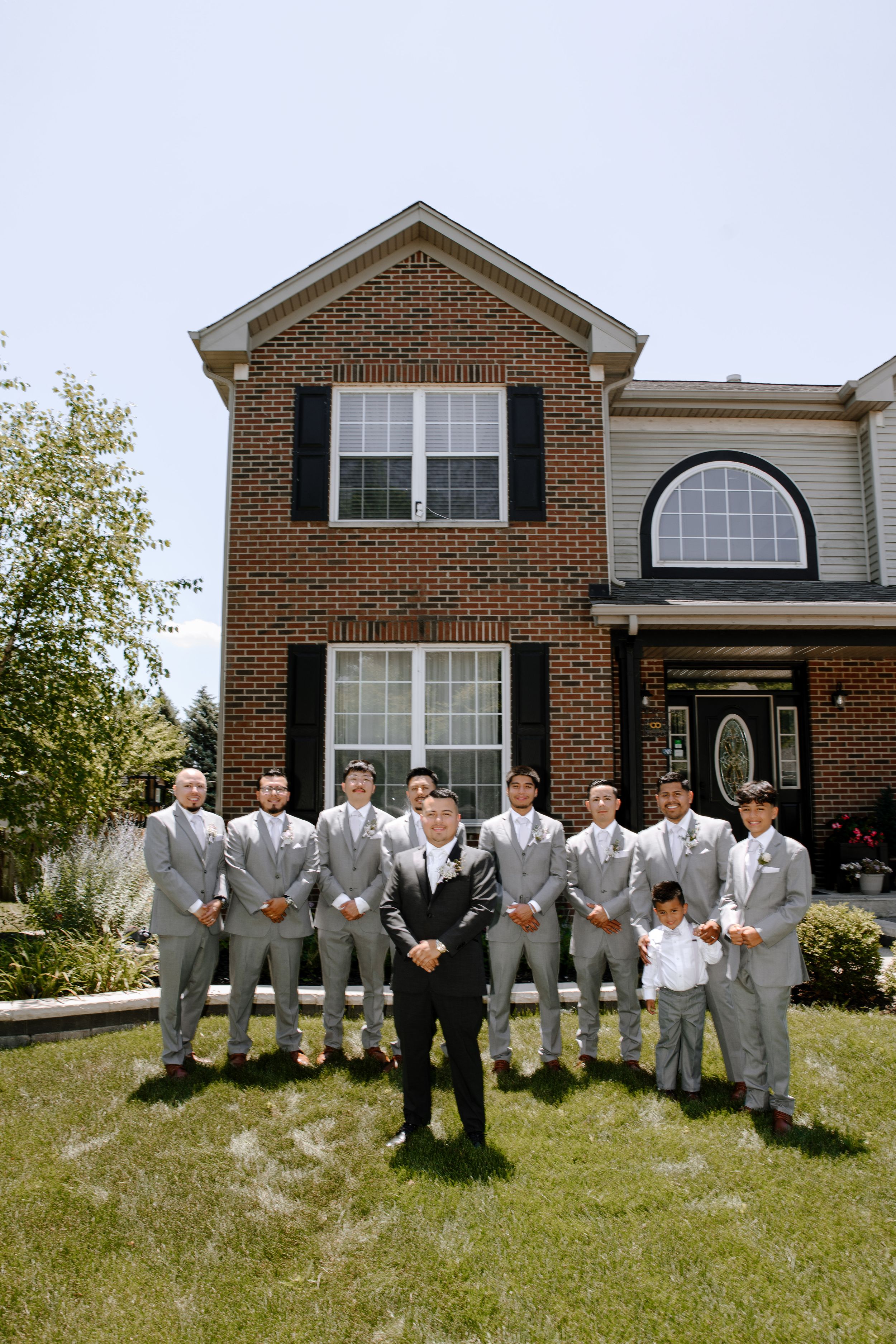 Groomsmen in light gray suits standing in front of a brick house on the lawn in varying poses.