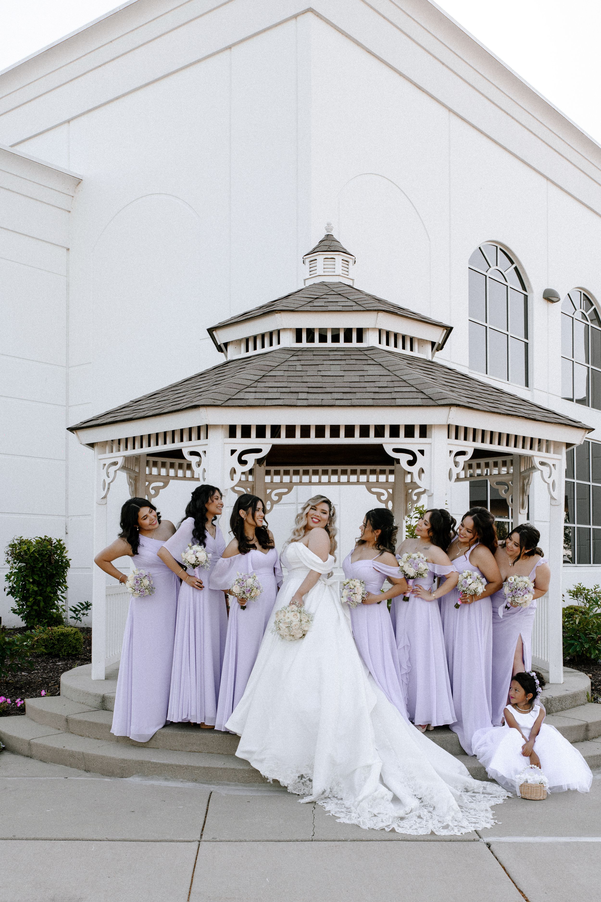 A group in lavender bridesmaids dresses and a white wedding gown pose together outside a white gazebo in playful poses.
