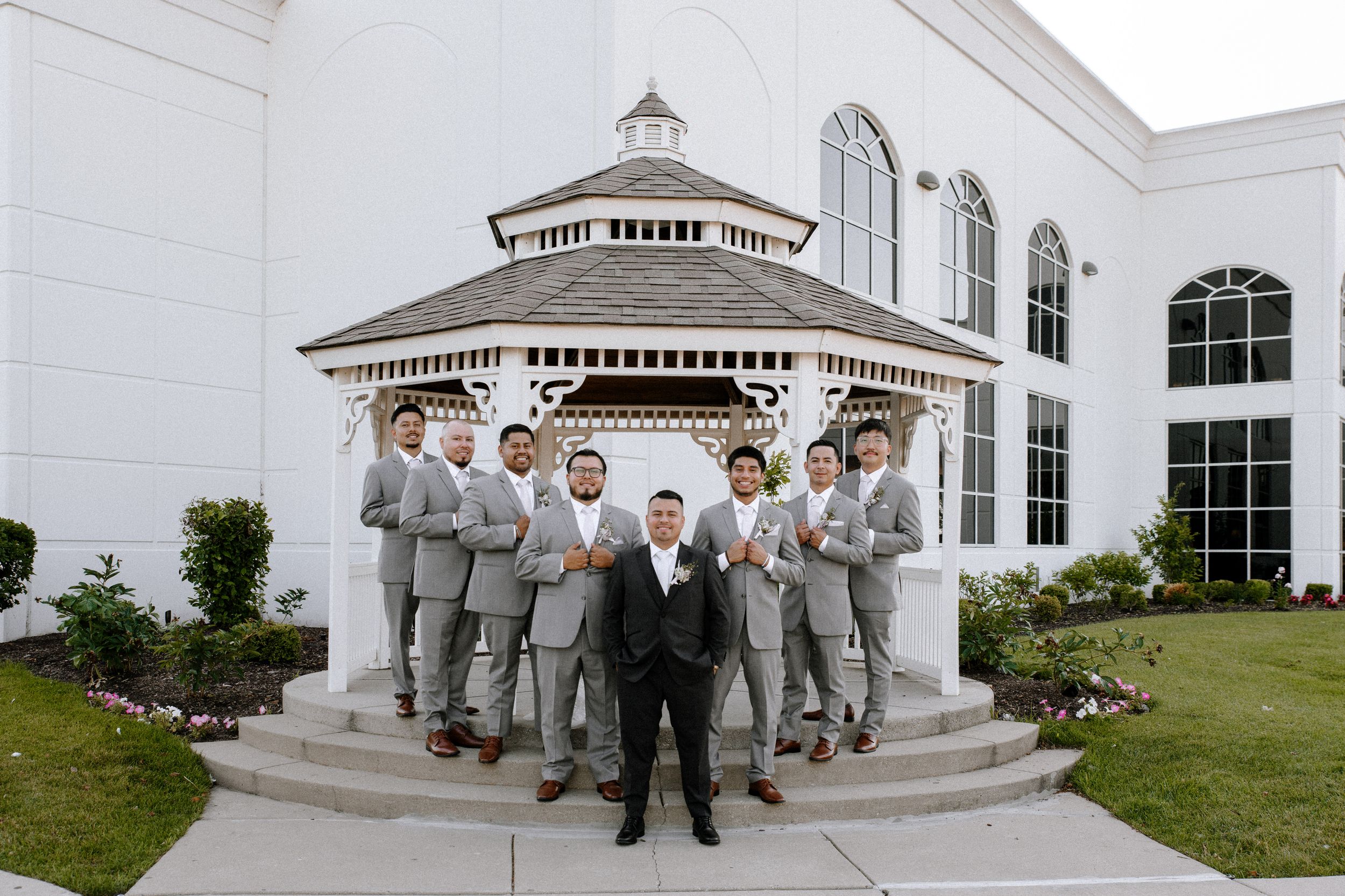 Wedding party poses for panoramic photos outside a white classical building with columns and arches.