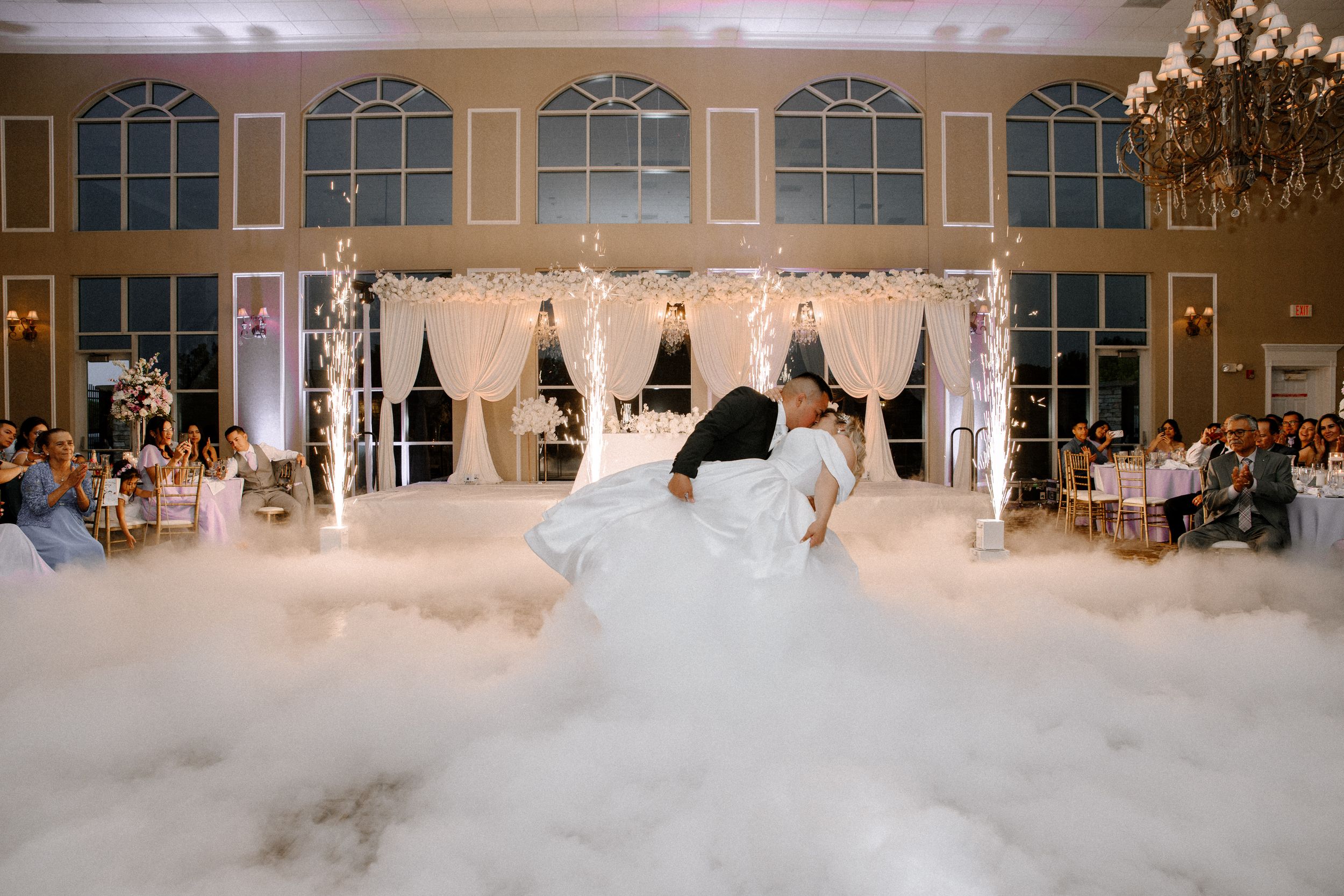 Elegant ballroom wedding venue illuminated by chandeliers with panoramic windows and couples dancing on the floor.