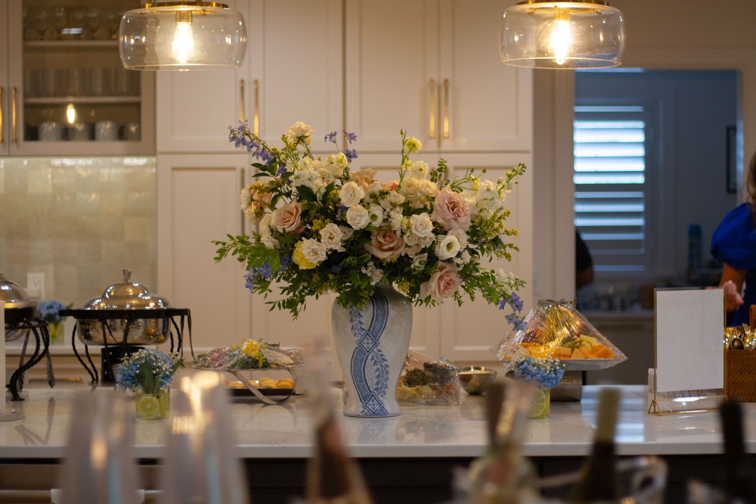 Elegant floral centerpiece with roses and greenery in a blue vase on a white kitchen counter with pendant lighting.