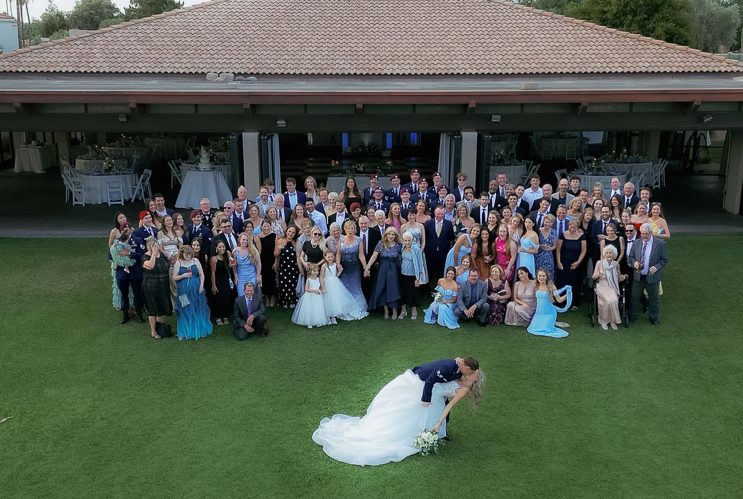 Large wedding party gathered on a green lawn in front of a covered porch with one couple posing dramatically in the foreground.