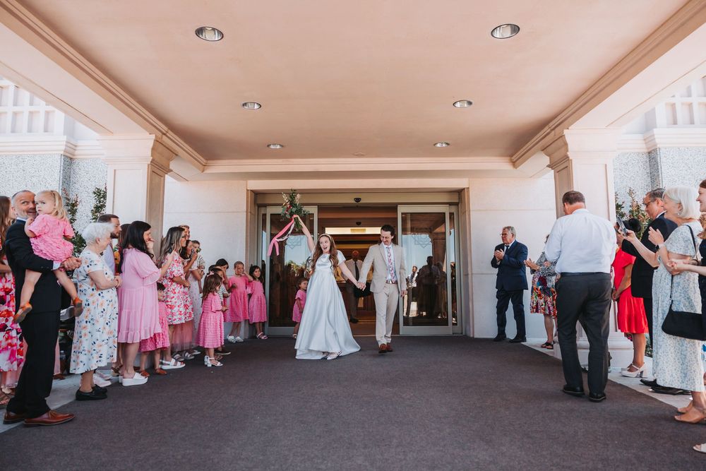 Wedding party exits church doors while guests line up on both sides with pink attire and colorful decorations.