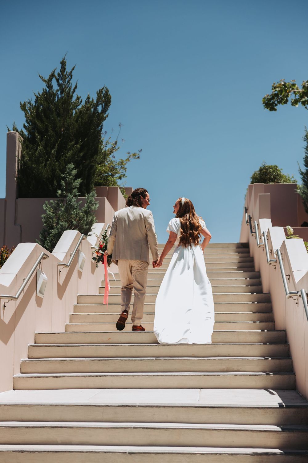 A couple climbs white concrete stairs on their wedding day, with the bride in a flowing white dress and groom in beige suit.