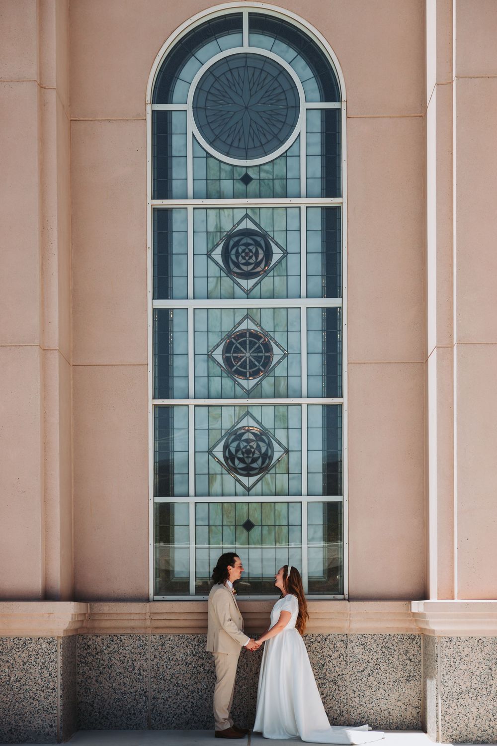 Two people stand together beneath a tall arched architectural window with ornate geometric patterns and designs.