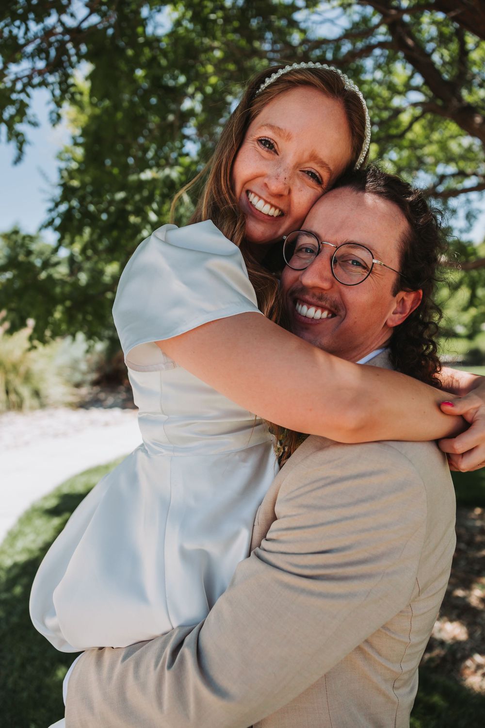 A joyful couple embraces outdoors on their wedding day, with one wearing a white dress and the other in a grey suit.