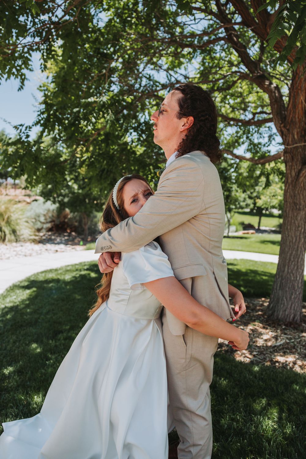 Couple embraces lovingly under shady green trees in an outdoor park setting during a summer afternoon.