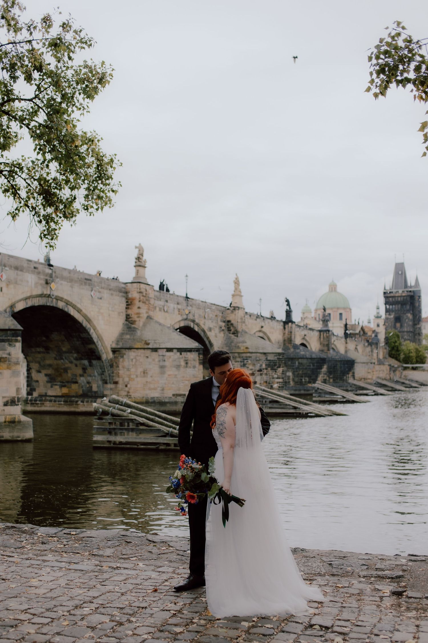 A couple in wedding attire share an intimate moment by Charles Bridge in Prague on a misty day.