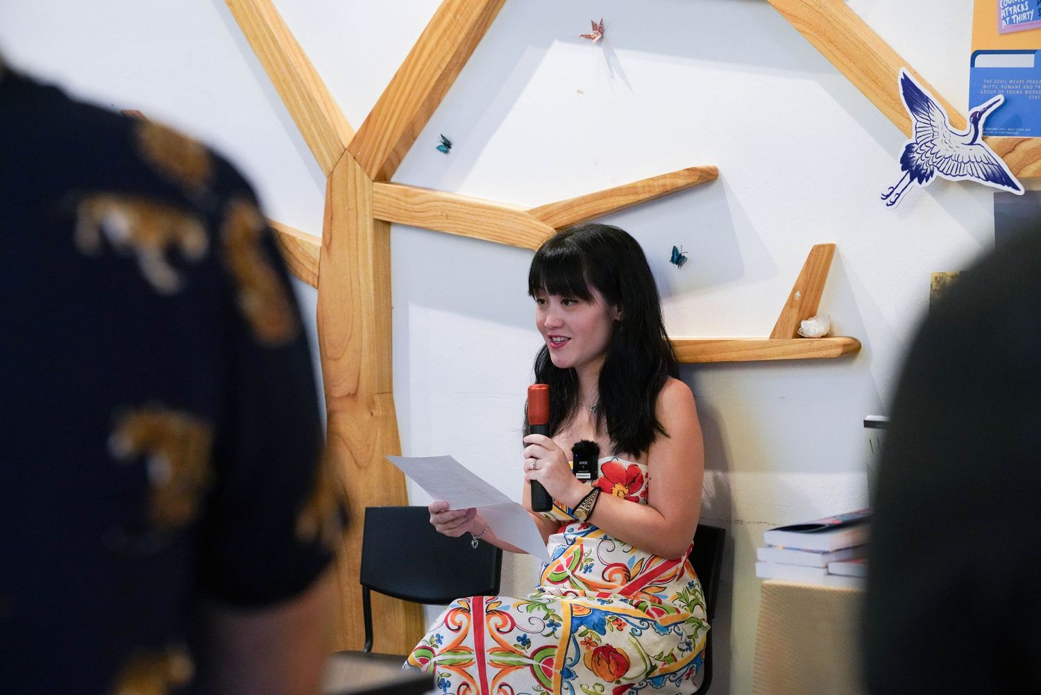 A person sitting in a wooden chair reads from papers during what appears to be a presentation or meeting.