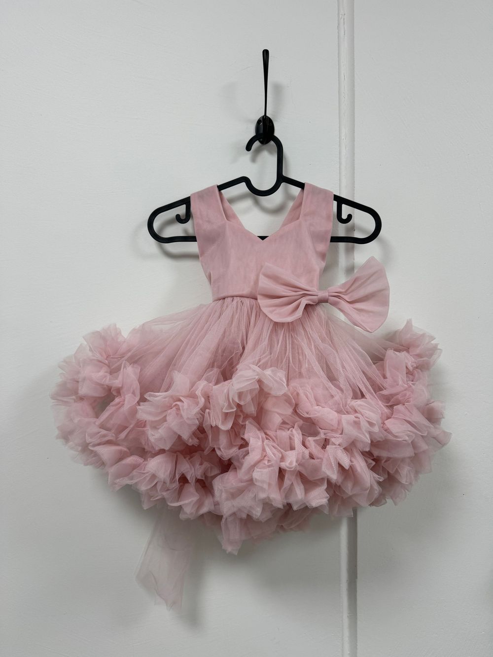 Pink tulle baby dress with large bow detail and ruffled skirt hanging on black hanger against white wall.