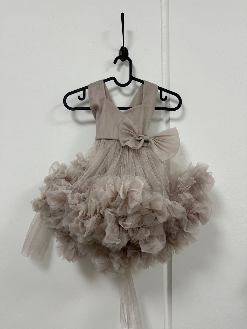 A delicate gray tulle dress with a ruffled tutu skirt hangs on a white wall displaying its fluffy layers and bow detail.