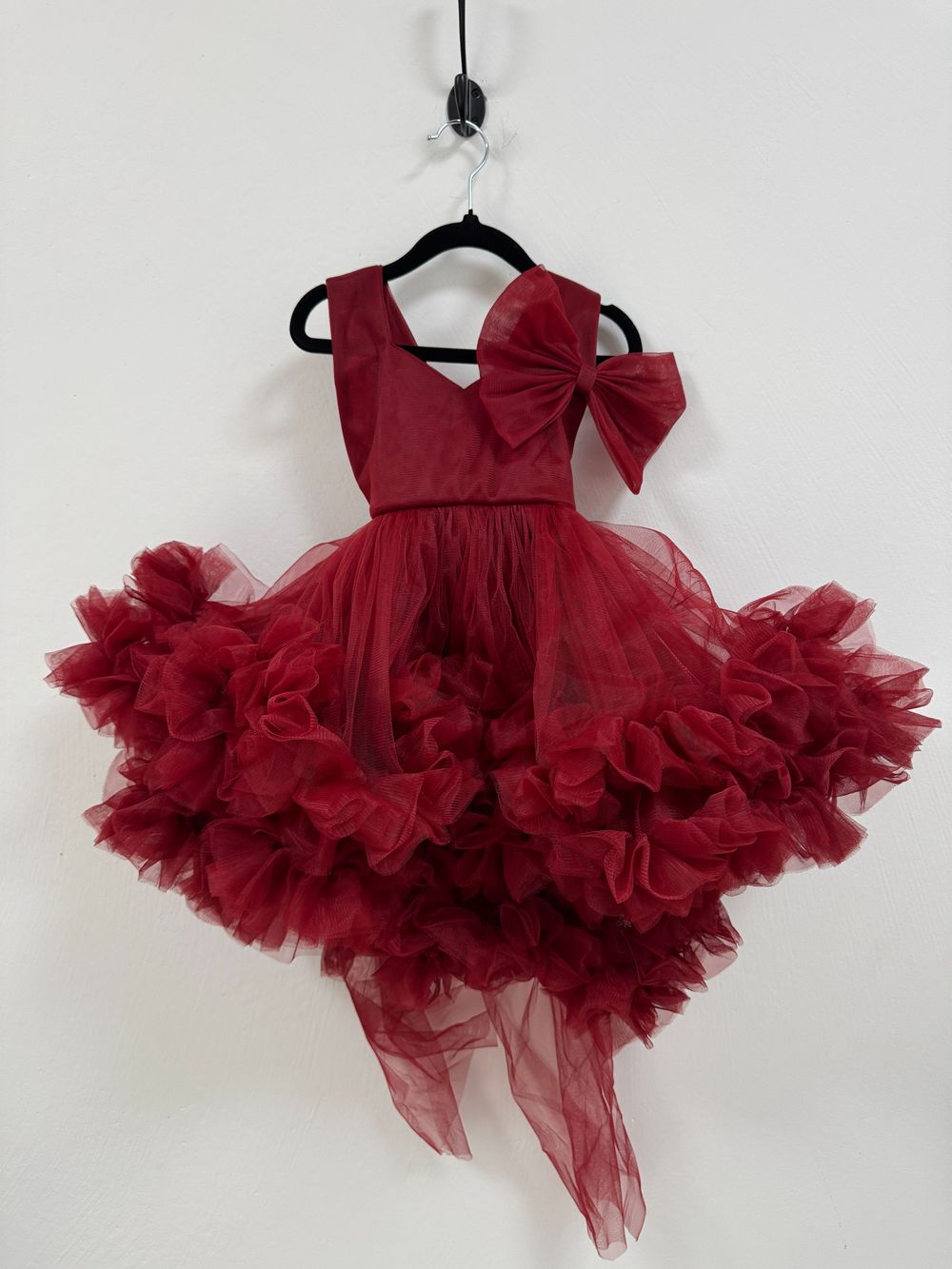 A burgundy tulle dress with ruffled layers and bow detail displayed from different angles on hangers.