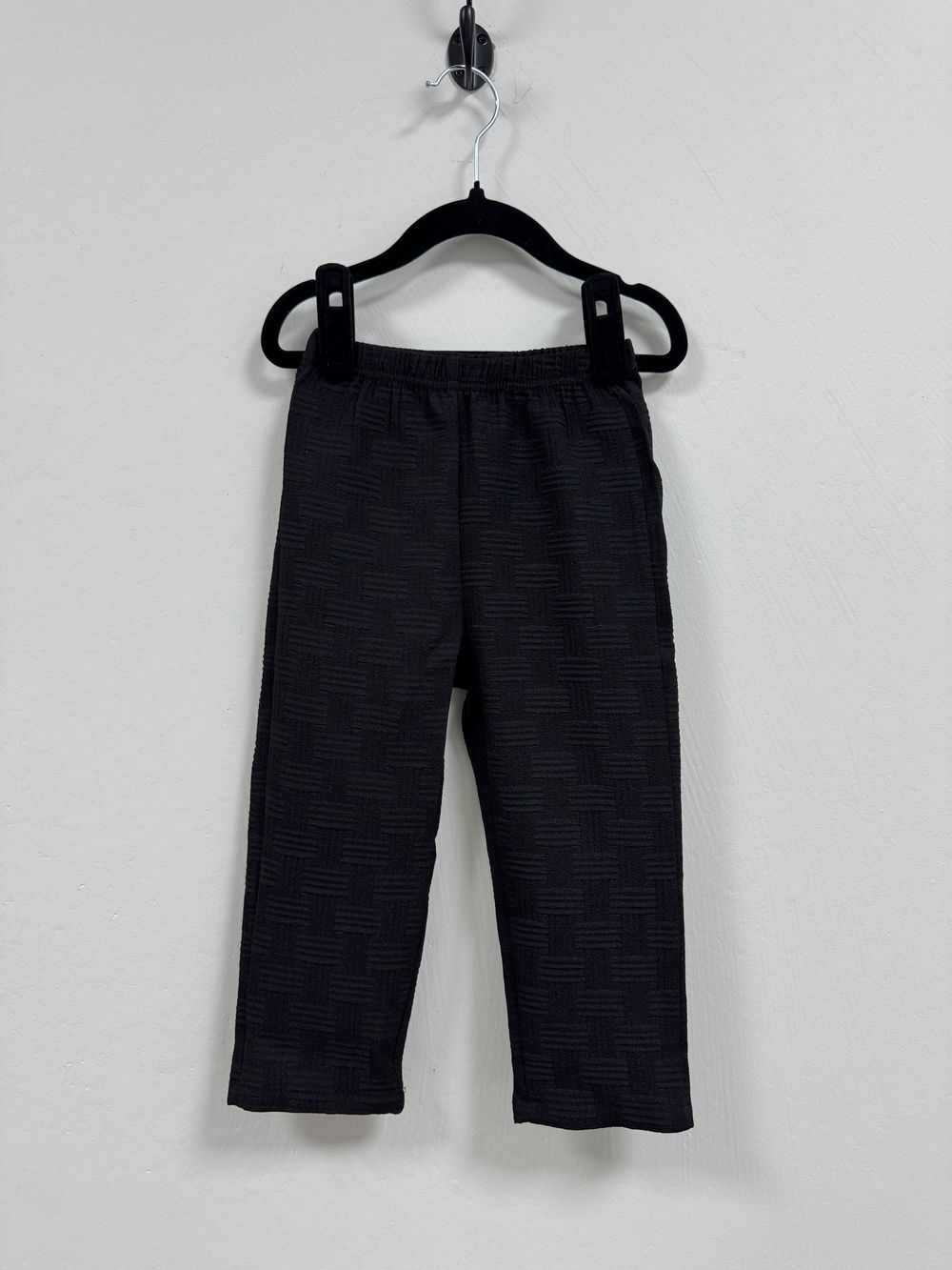 Black textured athletic pants hanging on plastic hanger against white wall.