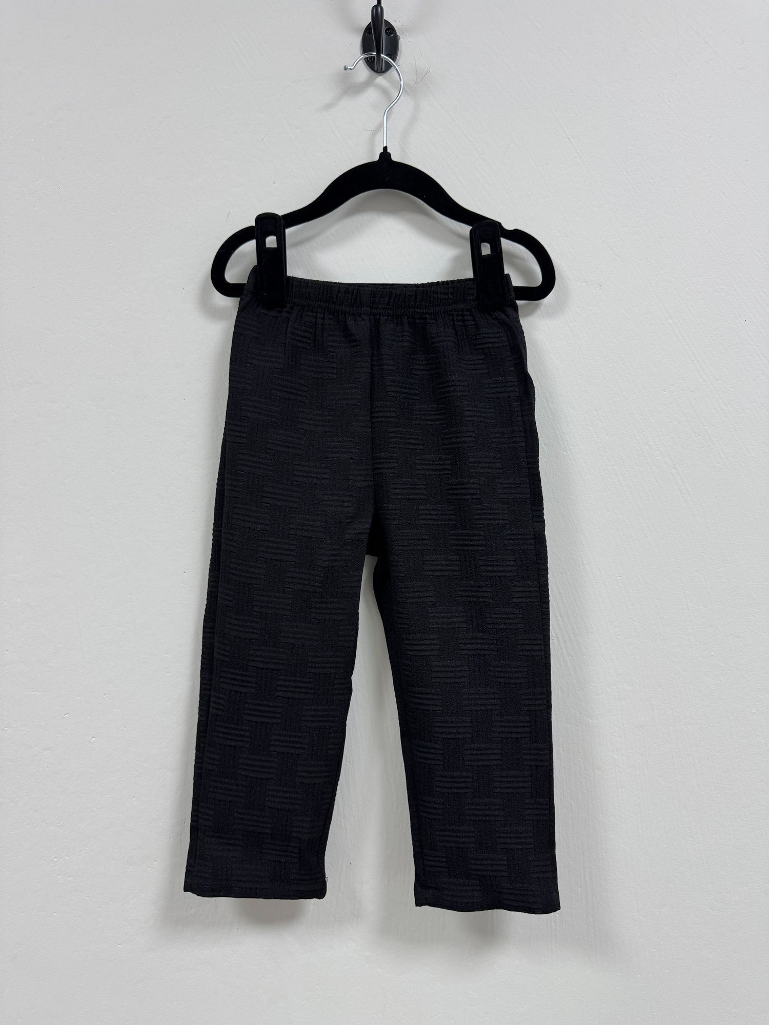 Black textured athletic pants hanging on plastic hanger against white wall.