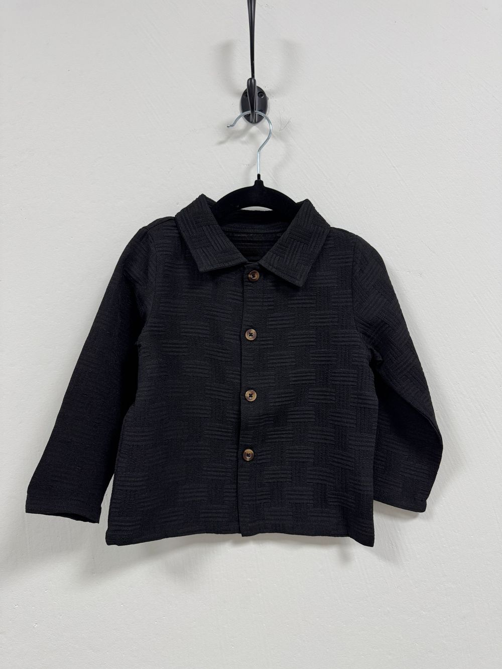 Black textured button-up jacket with collared neckline hanging on display against white background.