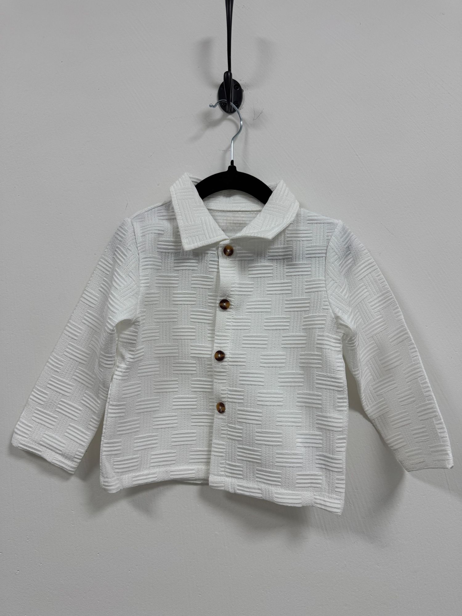 White textured long-sleeve button-up shirt for children hanging on black hanger against gray wall.