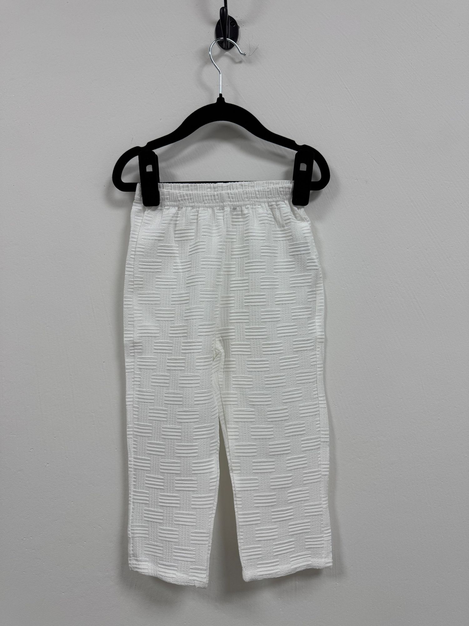 White textured elastic waist pants hanging on black plastic hanger against light gray wall.