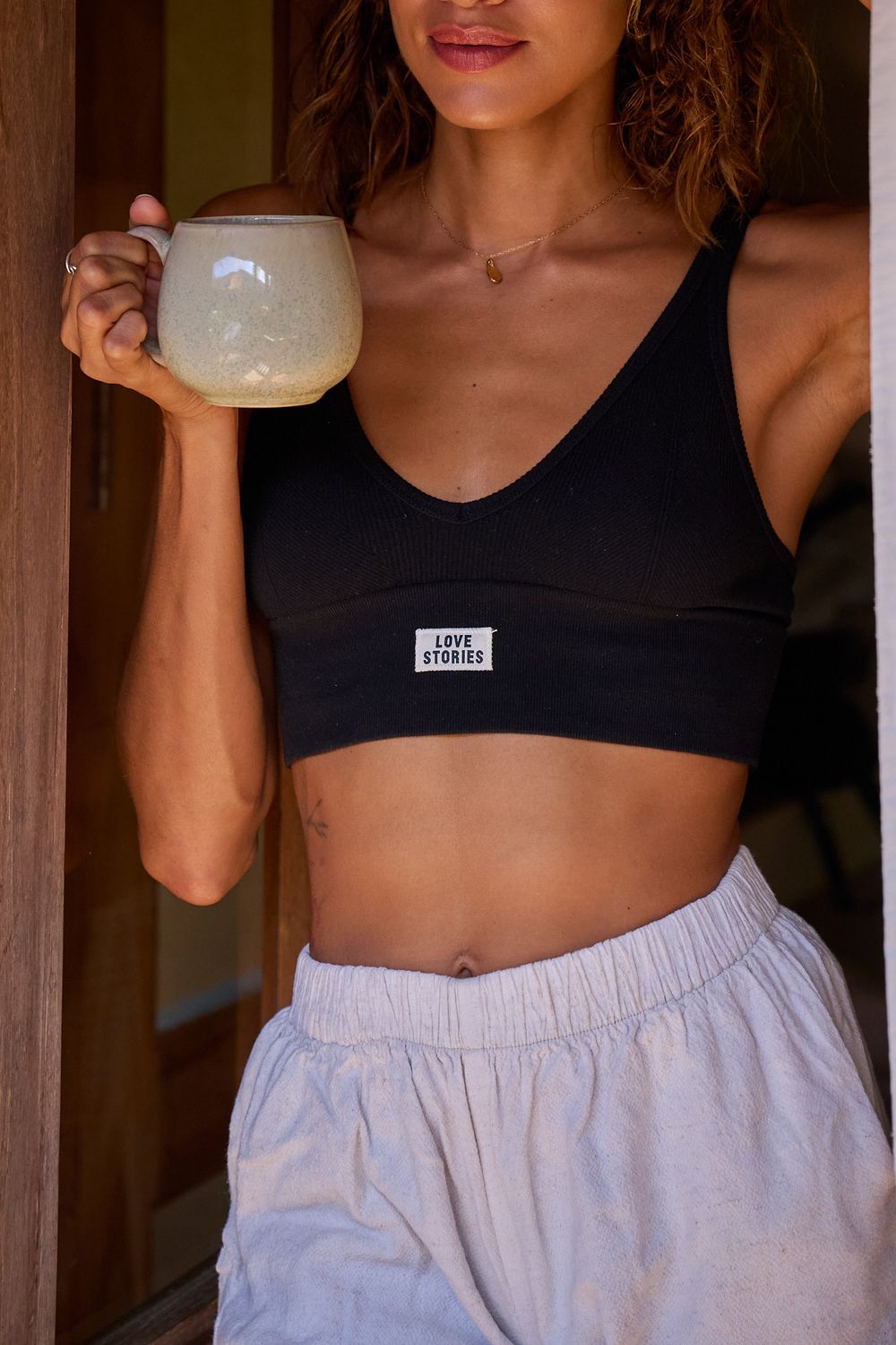 Person in black sports bra and white lounge shorts holds ceramic mug while standing in rustic doorway.