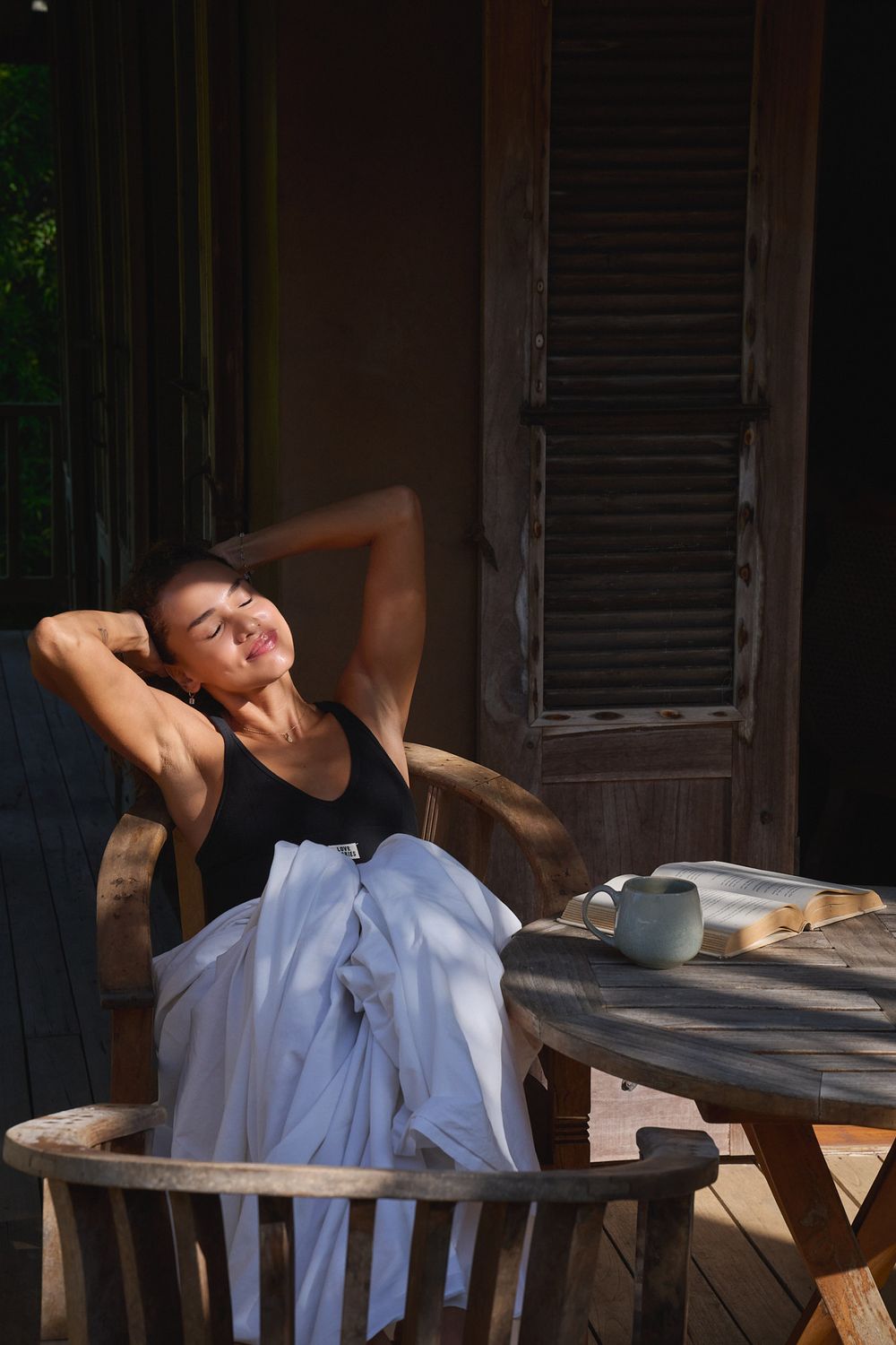 Relaxing figure in black tank top lounges on wooden porch chair beside table with tea and book in warm sunlight.