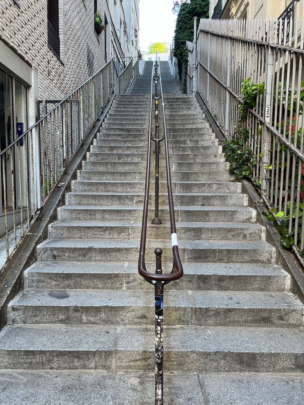 Long concrete staircase with central metal handrail leading upwards between buildings with a fence on one side.