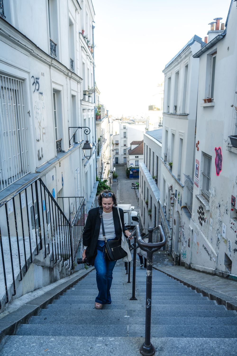 A person climbs steep stone steps in a narrow street of Montmartre, Paris with white buildings on both sides.