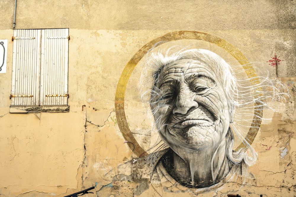 Street art mural with a golden halo on a weathered beige wall with window shutters visible.