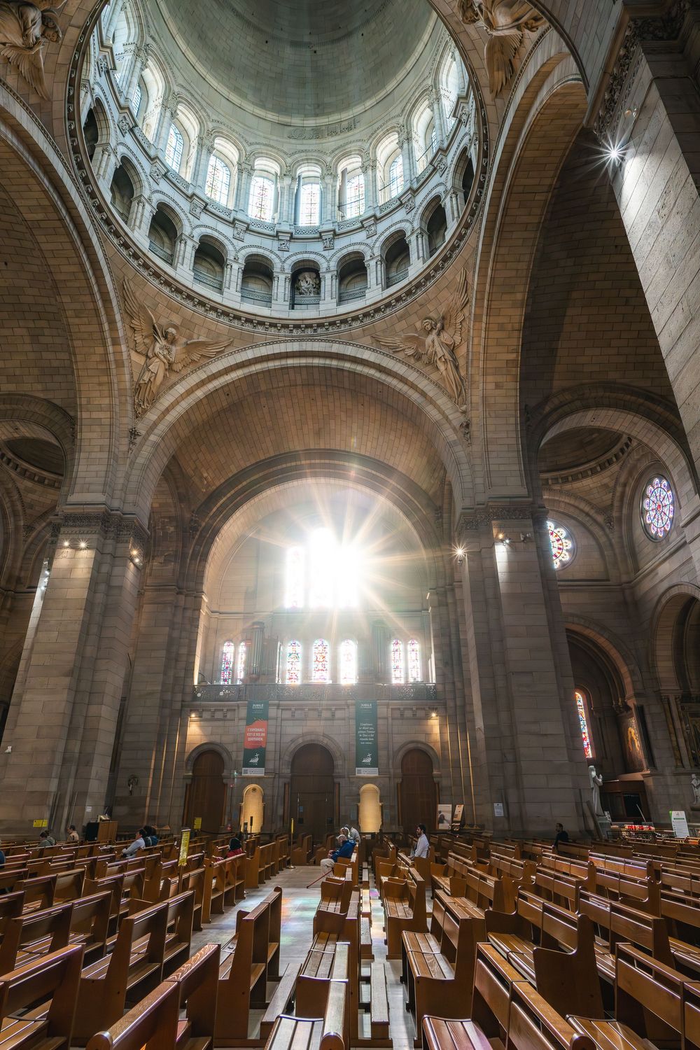 Sunlight streams through the windows of a grand cathedral interior with wooden pews and an impressive domed ceiling.