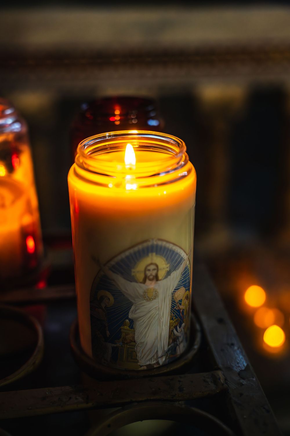 A lit religious prayer candle in a glass jar glows warmly in the darkness, casting a soft golden light.