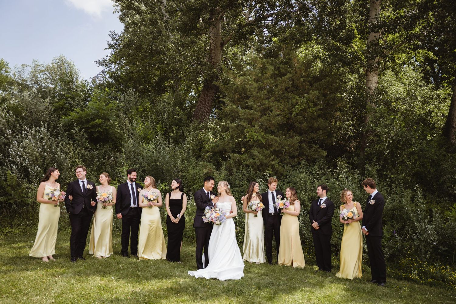 Wedding party in elegant attire stands in a line on green grass with lush trees and foliage in the background.